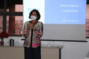 Professora Elizabeth Ribeiro, diretora da fundep, veste conjunto de blusa estampada em tons florais e saia preta. Ela tem cabelos pretos, é baixa, usa óculos e máscara branca e está gesticulando à frente da projeção da marca fundep concursos.